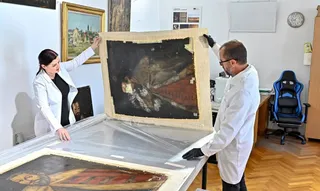 Două persoane în halate albe manipulează un tablou mare într-un atelier de restaurare, lângă alte tablouri și un scaun de birou.
