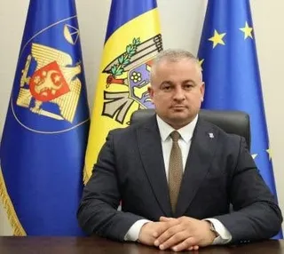 Un bărbat cu părul scurt și costum închis, purtând cămașă albă și cravată maro, stă la o masă în fața unor steaguri colorate.