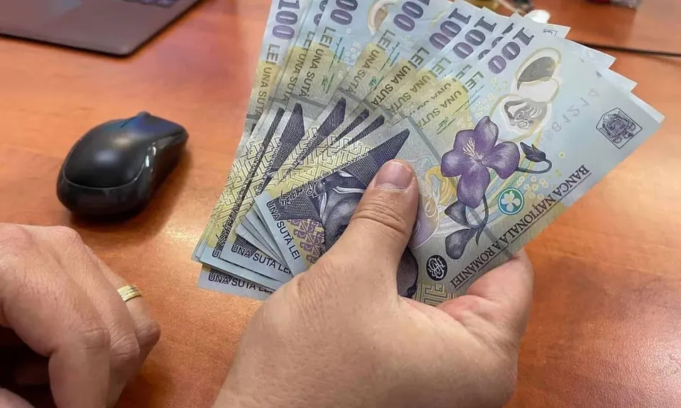 O persoană ține un teanc de bancnote de o sută de lei deasupra unei mese de lemn lângă o mouse de culoare neagră.