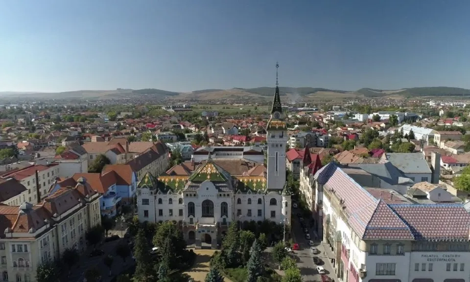 O vedere aeriană asupra unui oraș cu numeroase clădiri cu acoperișuri colorate și un turn înalt sub un cer senin.