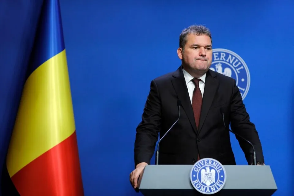 Un bărbat într-un costum negru și cravată roșie stă în spatele unui podium cu un emblema albastră în fața unui fundal albastru și a unui steag.