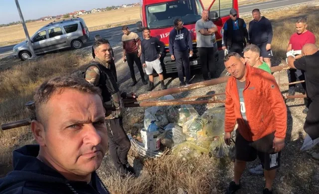 Un grup de bărbați stă pe un teren cu iarbă uscată lângă un camion roșu și saci cu deșeuri plini.