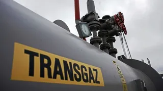 Un conducte gri mare cu inscripția TRANSGAZ pe un fundal de echipamente industriale și un cer încorporat.