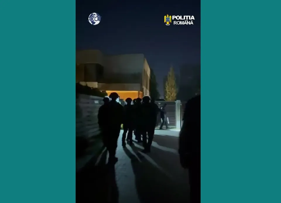 Siluete ale unor persoane în haine închise merg pe un drum lângă o clădire sub un cer nocturn.