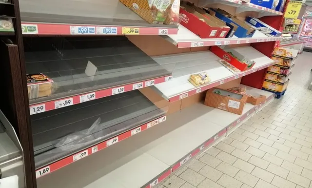 Rafturi de supermarket goale și parțial umplute cu câteva cutii de carton și produse ambalate sunt dispuse pe mai multe niveluri.