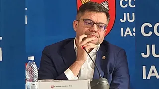 Un bărbat cu ochelari și sacou închis la culoare stă la o masă cu un microfon și o sticlă de apă în față.