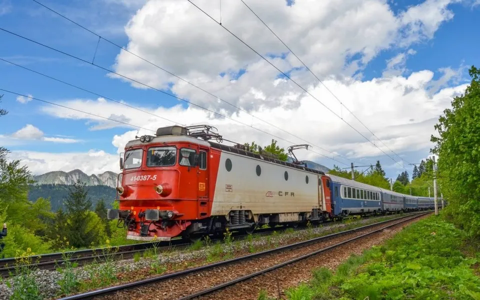 Un tren cu locomotivă roșie și albă tractează vagoane albastre pe o cale ferată sub un cer cu nori plutitori.
