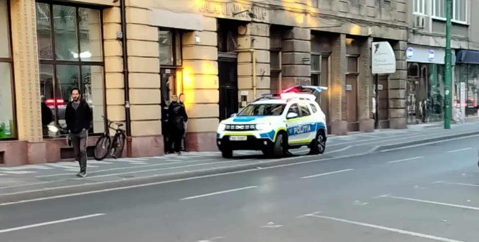 Un vehicul de poliție cu luminile aprinse este parcat pe marginea unei străzi lângă un trotuar și o clădire cu vitrine.