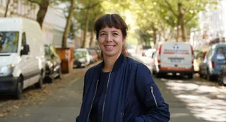 O femeie cu părul scurt și ciufulit poartă o jachetă albastră și zâmbește pe un drum urban cu mașini parcate și copaci în fundal.
