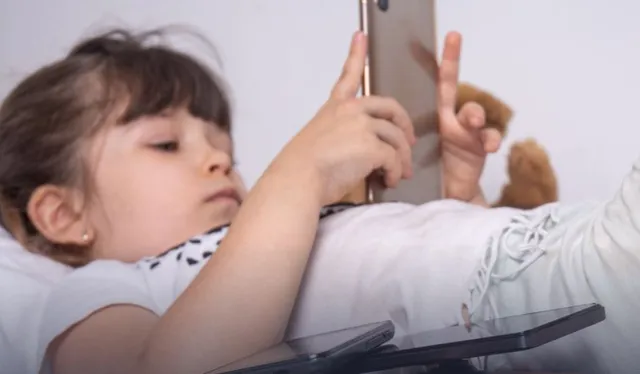 O copilă cu părul castaniu stă întinsă pe o suprafață albă, ținând un telefon mobil cu ambele mâini în fața feței.