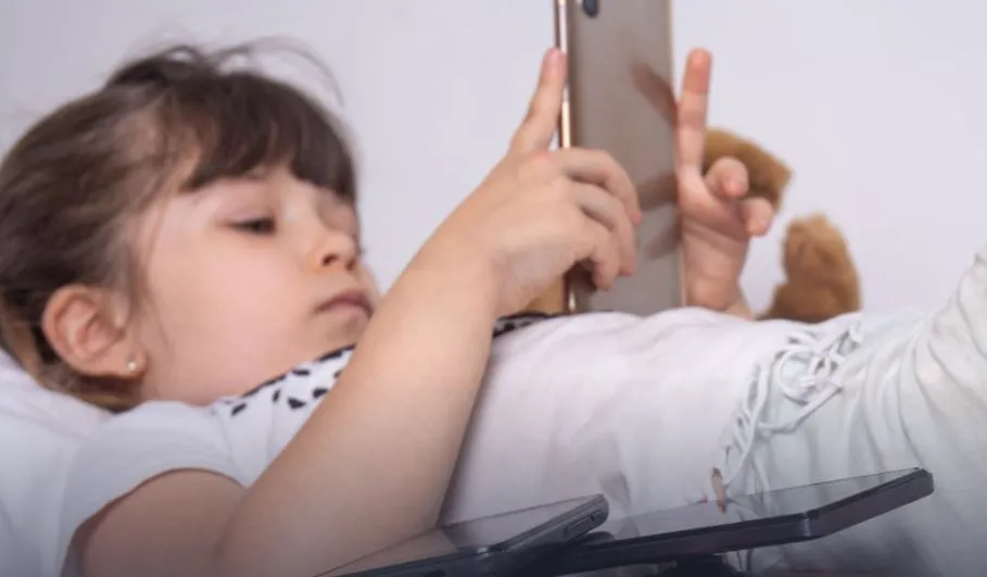 O copilă cu părul castaniu stă întinsă pe o suprafață albă, ținând un telefon mobil cu ambele mâini în fața feței.