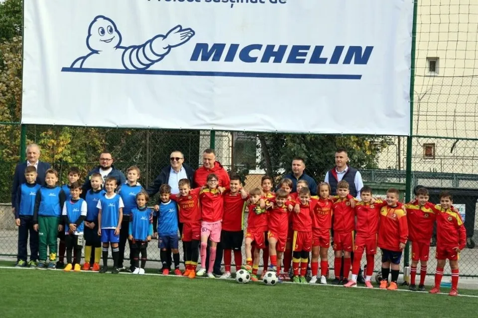 Un grup de copii în echipamente sportive roșii și albastre stau în rând pe un teren verde în fața unui banner alb mare.