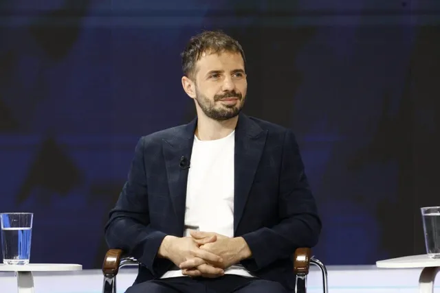 Un bărbat cu barbă, purtând un sacou închis la culoare peste un tricou alb, stă așezat cu mâinile încleștate în fața unui fundal albastru.