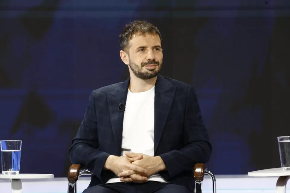 Un bărbat cu barbă, purtând un sacou închis la culoare peste un tricou alb, stă așezat cu mâinile încleștate în fața unui fundal albastru.