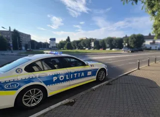 O mașină de poliție albă cu marcaje albastre și galbene este parcată pe marginea unui drum asfaltat lângă un trotuar din pavaj.