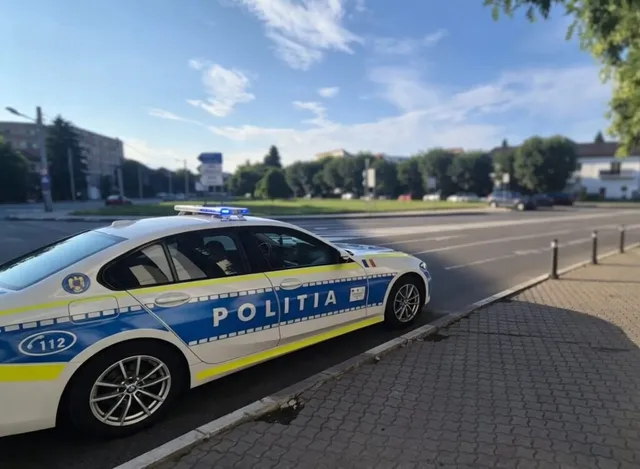 O mașină de poliție albă cu marcaje albastre și galbene este parcată pe marginea unui drum asfaltat lângă un trotuar din pavaj.