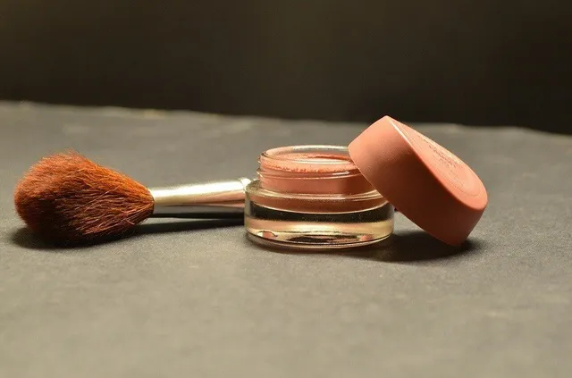 Un pensul de machiaj cu peri maro și un recipient mic de cosmetice deschis cu capac roz sunt așezate pe o suprafață gri închisă.
