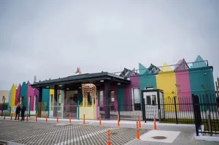 O clădire colorată cu panouri verticale galbene, roz și verzi este situată în spatele unui gard negru și a unor conuri portocalii.