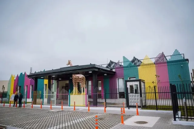 O clădire colorată cu panouri verticale galbene, roz și verzi este situată în spatele unui gard negru și a unor conuri portocalii.