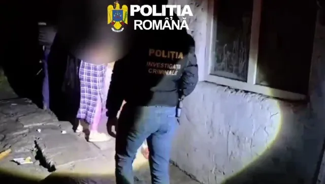 Un agent de poliție cu inscripție pe spate stă lângă un perete al unei clădiri lângă o persoană purtând pantaloni cu model pătratat.