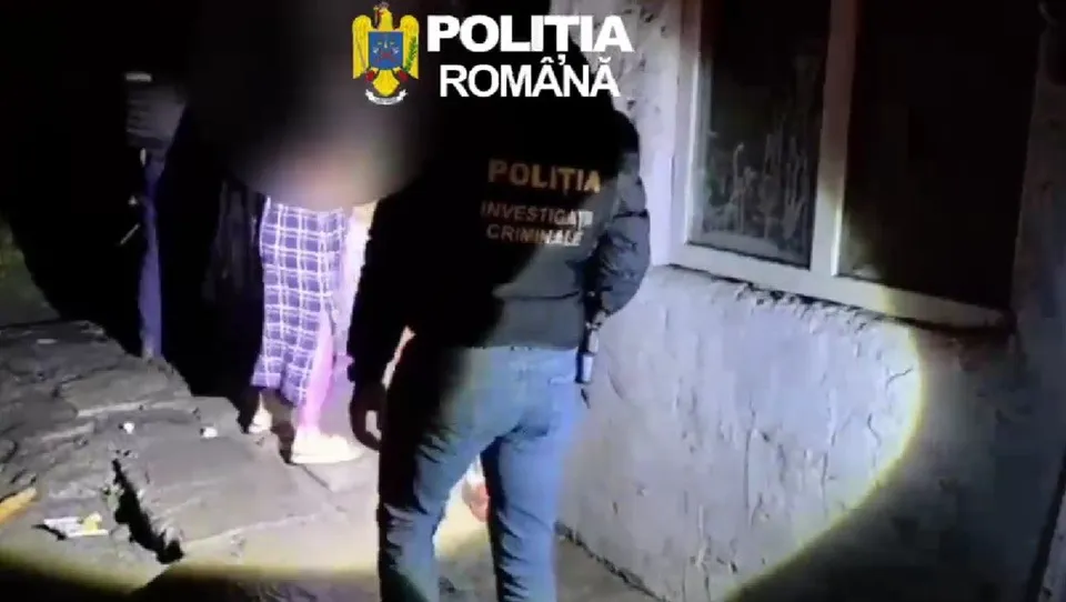 Un agent de poliție cu inscripție pe spate stă lângă un perete al unei clădiri lângă o persoană purtând pantaloni cu model pătratat.