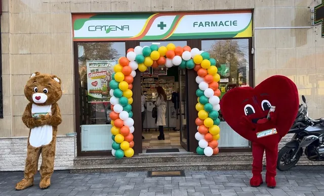 Două mascote, una de urs și una în formă de inimă, stau pe trotuar lângă o intrare de farmacie decorată cu un arc de baloane colorate.