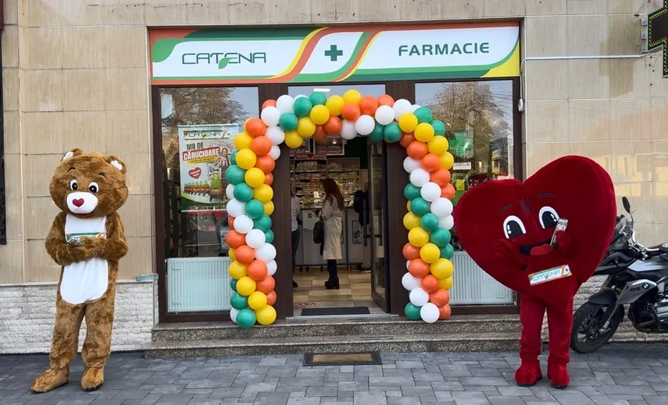 Două mascote, una de urs și una în formă de inimă, stau pe trotuar lângă o intrare de farmacie decorată cu un arc de baloane colorate.