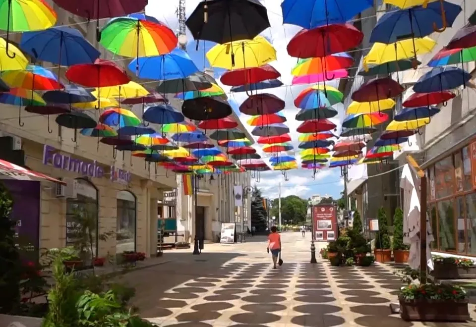 Multe umbrele colorate sunt suspendate deasupra unei străzi pietonale cu clădiri și plante în fundal.
