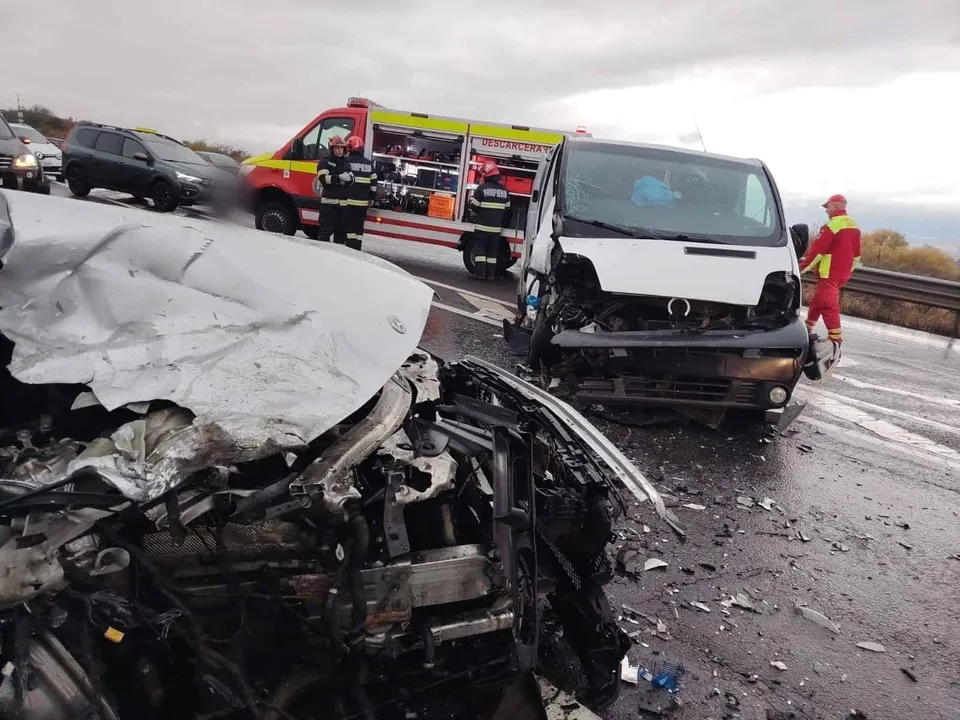 Un autoturism alb zdrobit și o autoutilitară albă sunt situate pe un drum umed lângă un vehicul de serviciu roșu și galben.