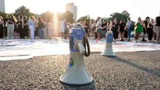 Un megafon alb cu mâner albastru stă pe un asfalt umed în fața unui grup de persoane aflate în lumina soarelui.