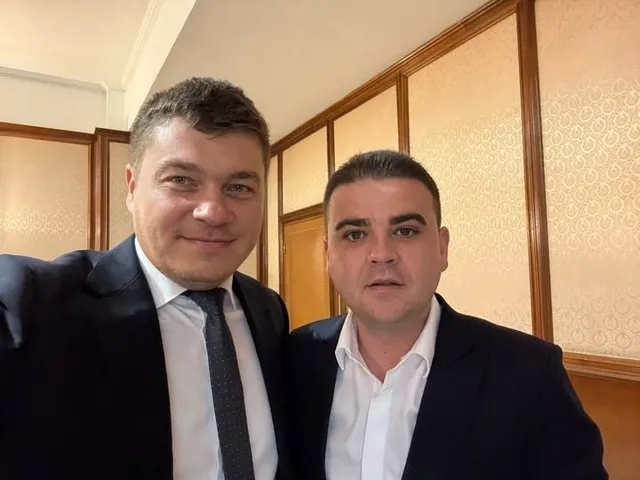 A doi bărbați îmbrăcați în costume și cămași albe stau unul lângă altul în fața unui perete cu panouri de lemn.