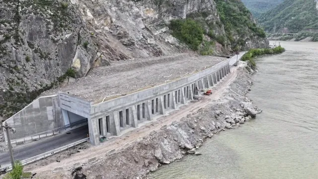 O structură de beton cu piloni se află pe malul unui râu, lângă o intrare de tunel și un drum asfaltat.
