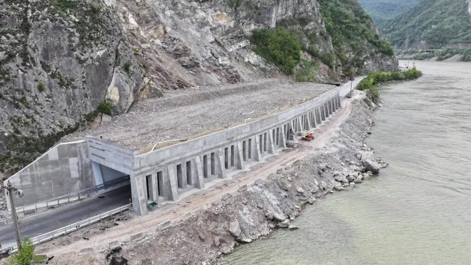 O structură de beton cu piloni se află pe malul unui râu, lângă o intrare de tunel și un drum asfaltat.