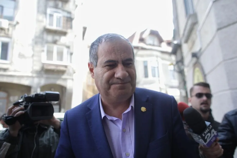 Un bărbat cu sacou albastru și cămașă lila stă în fața unor microfoane și a unei camere video în fața unor clădiri.