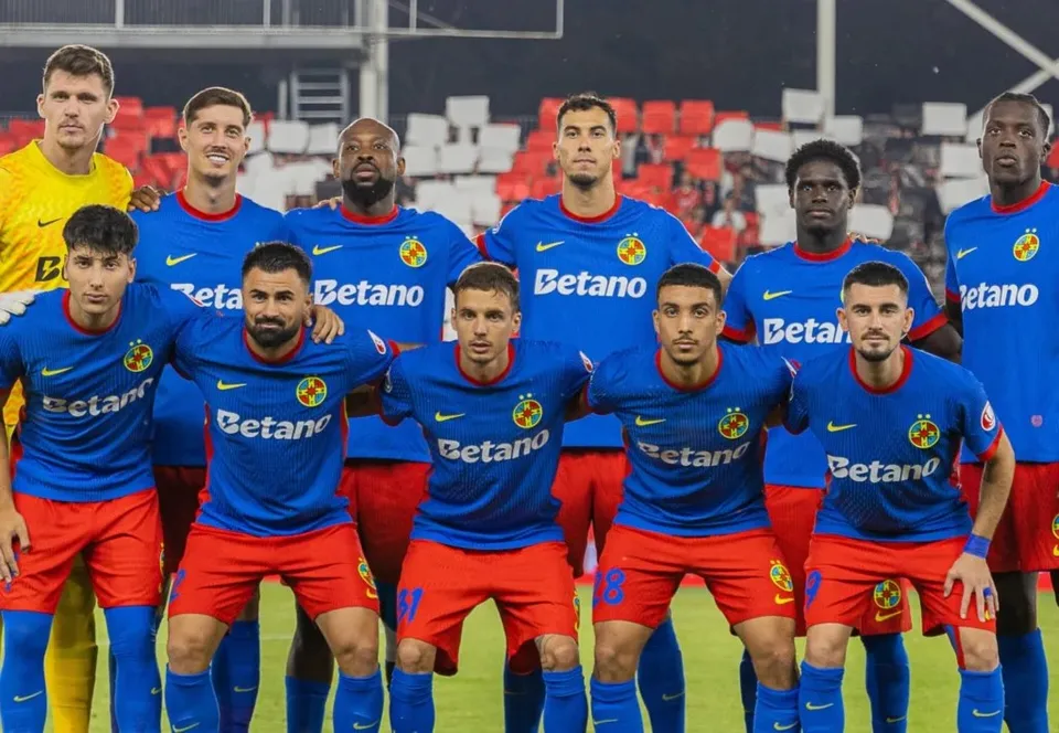 Un grup de fotbaliști în echipamente sportive albastre și roșii pozează într-o poziție de echipă pe un teren de fotbal.