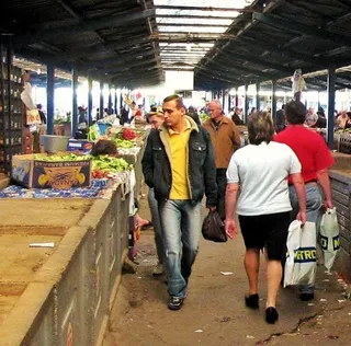 Un bărbat cu sac de cumpărături merge pe un culoar de piață printre alți oameni și tarabe cu fructe și legume sub un acoperiș de lemn.