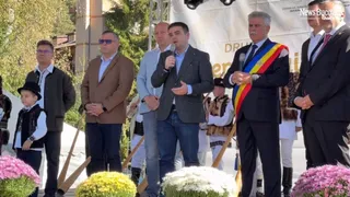 Un grup de bărbați stă în picioare în fața unui panou alb, în timp ce unul dintre ei ține un microfon.