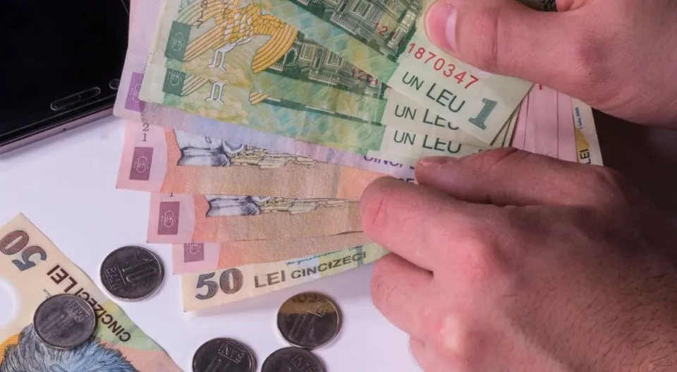 Două mâini țin un teanc de bancnote de lei și câteva monede pe o suprafață albă.