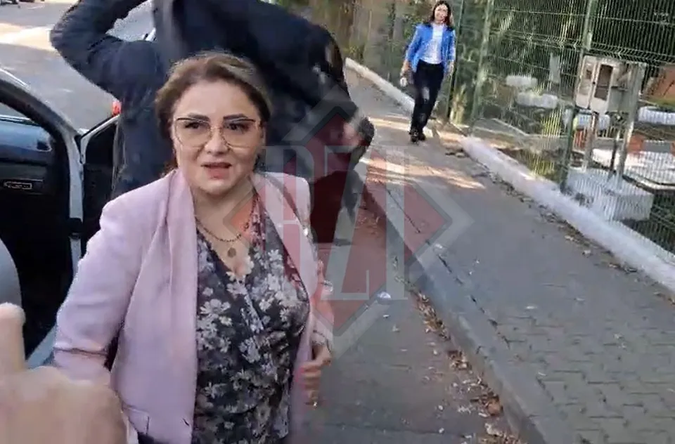 O femeie cu ochelari, purtând o sacou roz și o bluză înflorită, merge pe lângă o mașină deschisă pe un trotuar.