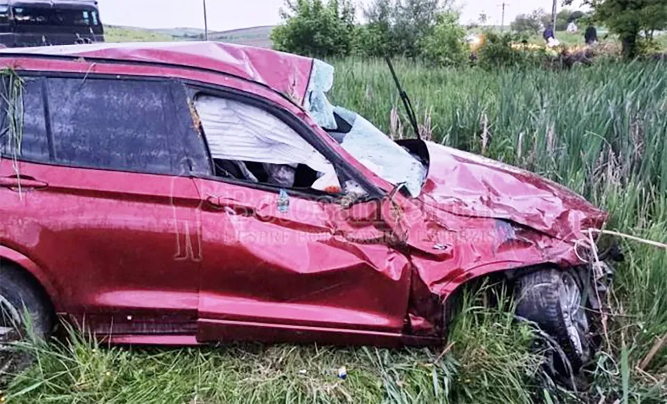 Un autoturism roșu este situat într-o zonă cu vegetație înaltă, prezentând avarii majore la nivelul capotei și portierei laterale.
