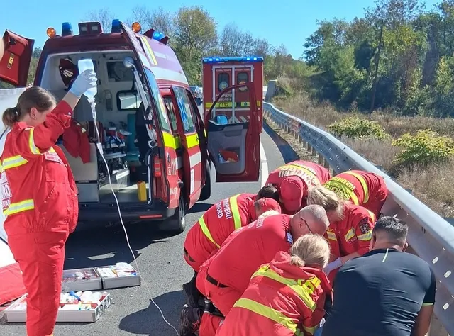 Echipe de intervenție în uniforme roșii oferă asistență medicală pe marginea unui drum, lângă două ambulanțe parcate pe o porțiune de asfalt.