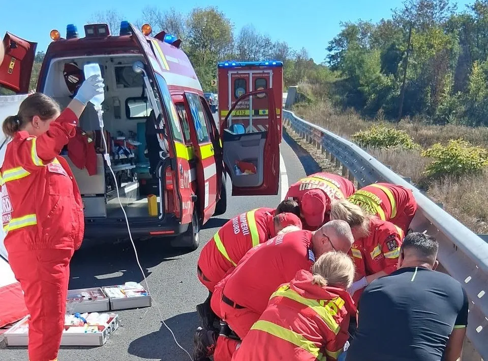 Echipe de intervenție în uniforme roșii oferă asistență medicală pe marginea unui drum, lângă două ambulanțe parcate pe o porțiune de asfalt.