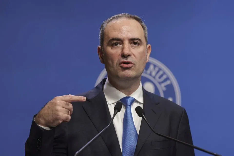 Un bărbat cu părul scurt, purtând sacou închis la culoare, cămașă albă și cravată albastră, stă în fața a două microfoane pe un fundal albastru.