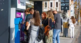 Un grup de persoane stă la un aparat de vânzare a biletelor de lângă un autobuz într-un mediu urban.