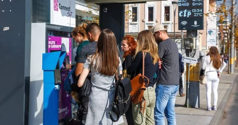 Un grup de persoane stă la un aparat de vânzare a biletelor de lângă un autobuz într-un mediu urban.