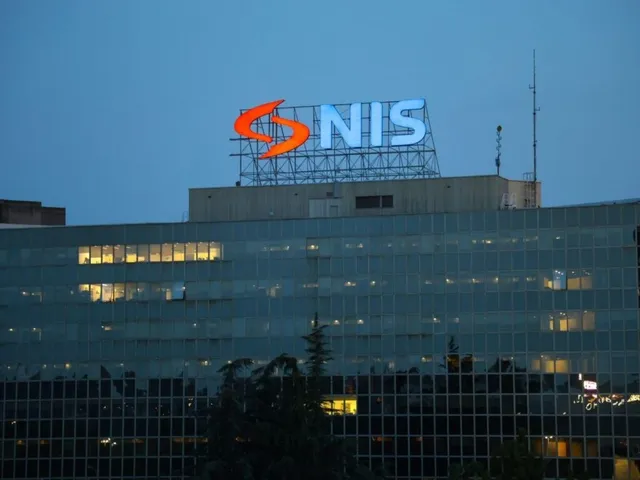 Un semn luminos cu textul NIS și un simbol portocaliu este montat pe acoperișul unei clădiri de birouri cu ferestre iluminat.