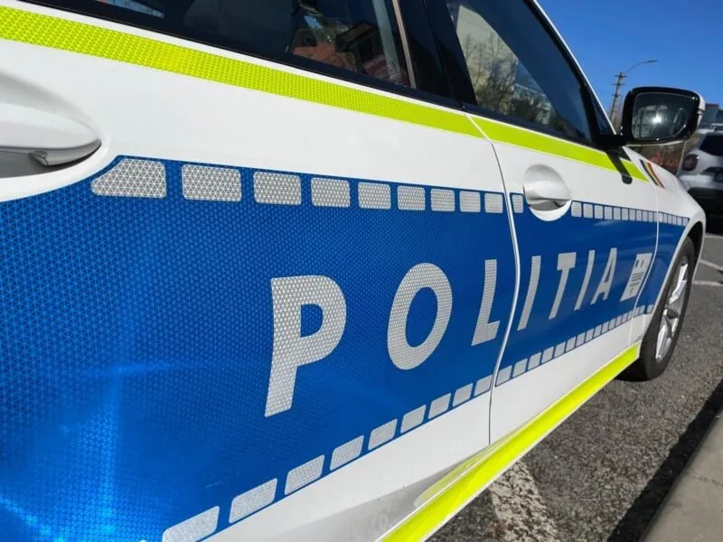 O parte laterală a unei mașini albe cu inscripția POLITIA scrisă cu litere gri pe un fundal albastru și benzi galbene reflectorizante.