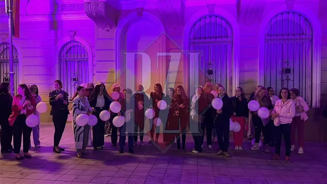 Un grup de persoane stă în fața unei clădiri iluminate în roz, ținând baloane albe.