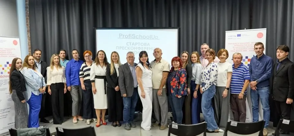 Un grup de oameni stă în rând în fața unui ecran de proiecție într-o sală cu perdele negre.