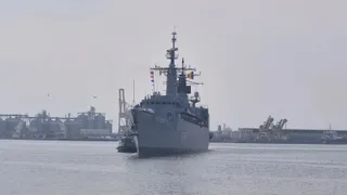 O navă militară gri navighează pe apă, având structuri portuare și industriale în fundal sub un cer înnorat.